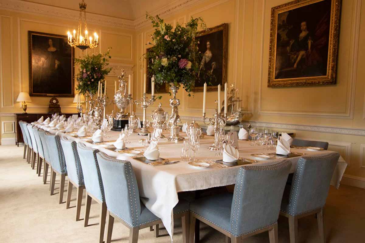 A dining room set for a beautiful and elegant corporate event lunch with long rectangular table and blue chairs.  The table is elaborately dressed with silver, fresh flower arrangements, candles and napkins.