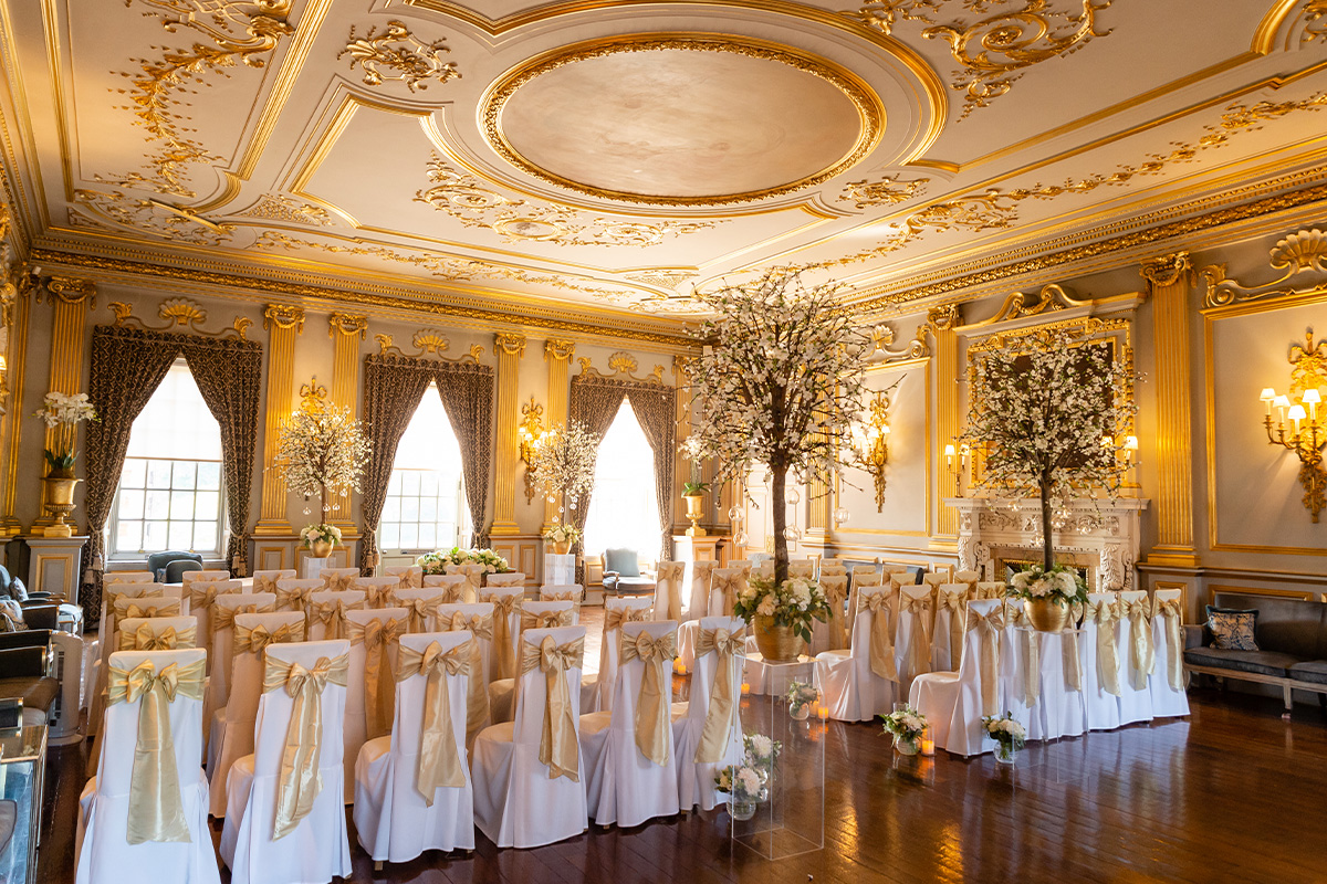 White chairs with golden ribbon draped down the back in a golden embellished room with white trees.  Dressed for a wedding ceremony at Knowsley Hall.