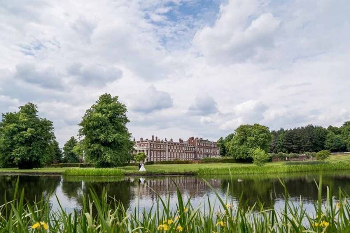 A bride & groom pictured standing in beautiful gardens by a lake and a stately home behind them